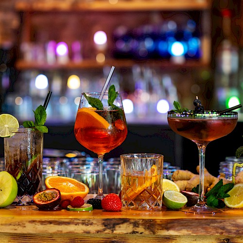 An array of colorful cocktails with garnishes like lime, mint, and berries on a bar counter, surrounded by sliced fruits and herbs.
