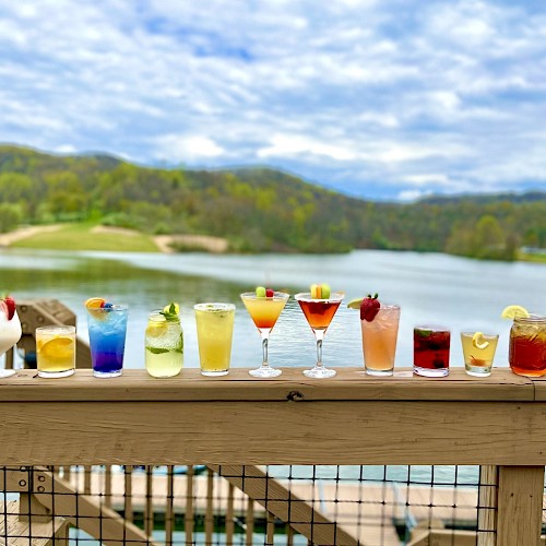 A row of colorful cocktails on a wooden railing, with a scenic lake and hills in the background under a cloudy sky.