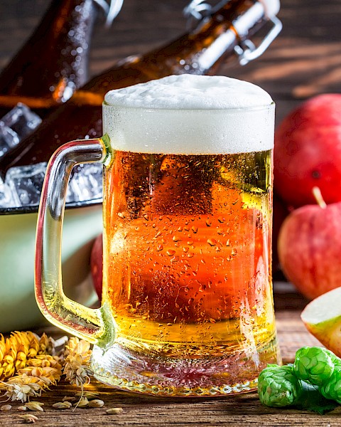 A glass of beer with a foamy head, bottles in ice, apples, wheat, and hops arranged on a wooden surface complete the scene.