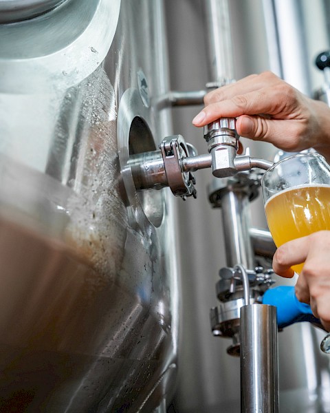 Someone filling a glass with beer from a large stainless steel brewing tank in a brewery setting.