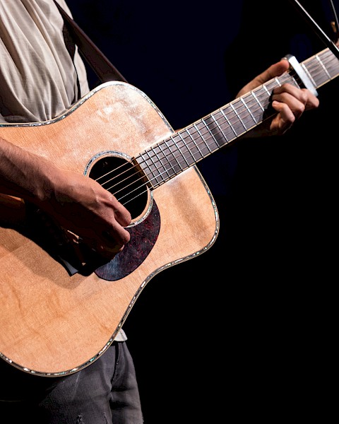 A person is playing an acoustic guitar on stage, holding a pick while wearing a casual shirt, with a microphone visible in the background.