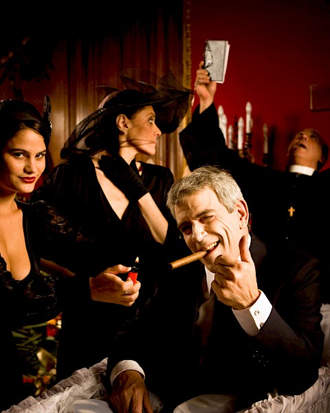 A man in a coffin with a cigar, surrounded by people, some in dramatic poses, in a dimly lit room.