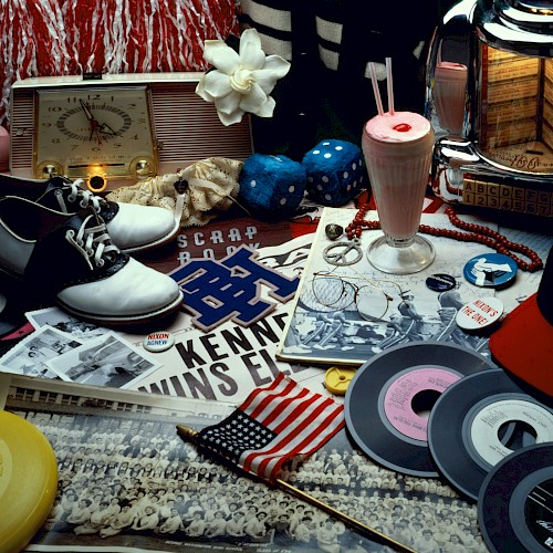 The image shows 1950s memorabilia, including records, saddle shoes, a milkshake, vintage photos, and an American flag.