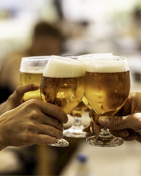 Four people clinking glasses of beer in a celebratory toast, with a blurred background indicating an indoor social gathering.