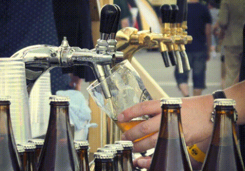 A person is pouring beer from a tap into a glass, surrounded by bottles and cups at an outdoor setting with multiple taps visible.