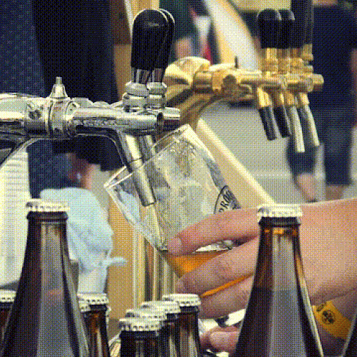 A person is pouring beer from a tap into a glass, surrounded by bottles and cups at an outdoor setting with multiple taps visible.