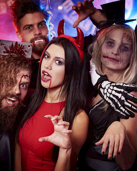 A group of people in Halloween costumes, including a witch, vampire, and skeleton, posing playfully with decor in the background.