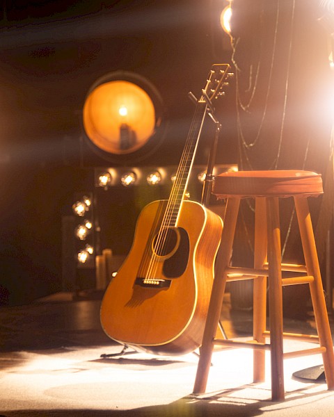 A guitar rests against a wooden stool on stage, surrounded by bright spotlights, creating a warm, inviting atmosphere.