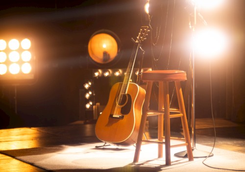 A guitar rests against a wooden stool on stage, surrounded by bright spotlights, creating a warm, inviting atmosphere.