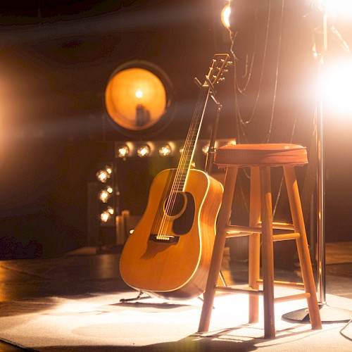 A guitar rests against a wooden stool on stage, surrounded by bright spotlights, creating a warm, inviting atmosphere.