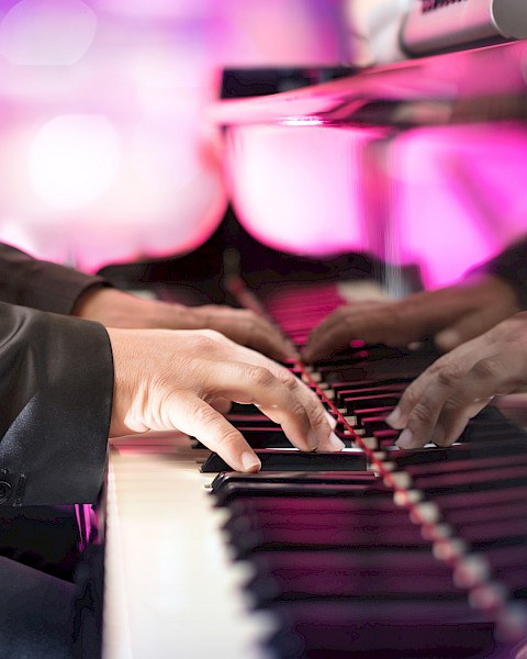 A person in a suit playing a grand piano, with vibrant pink and purple lighting reflecting off the polished surface.