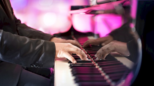 A person in a suit playing a grand piano, with vibrant pink and purple lighting reflecting off the polished surface.
