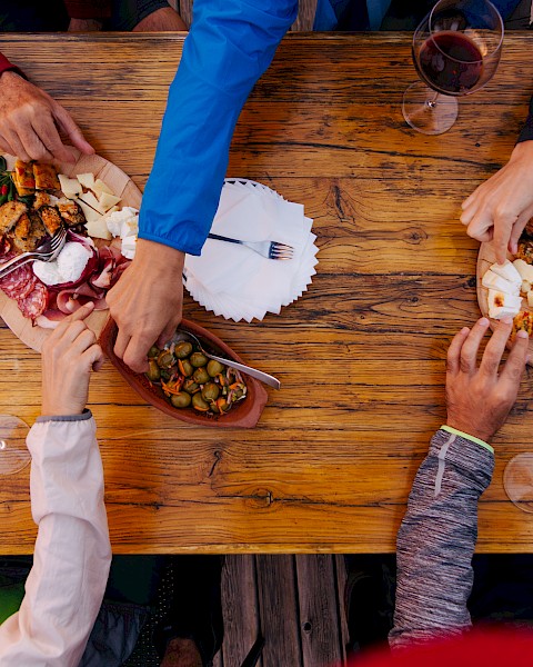 A group of people sharing charcuterie boards and wine at a wooden table, with various meats, cheeses, and bread.