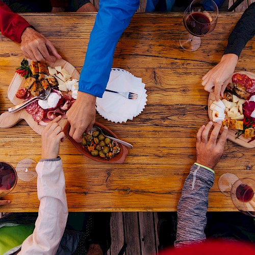 A group of people sharing charcuterie boards and wine at a wooden table, with various meats, cheeses, and bread.