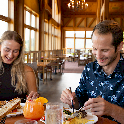 A couple enjoying a bright restaurant meal: woman with wine, man cutting a dish, sunny windowed lodge-style dining, tasty food and orange drinks.