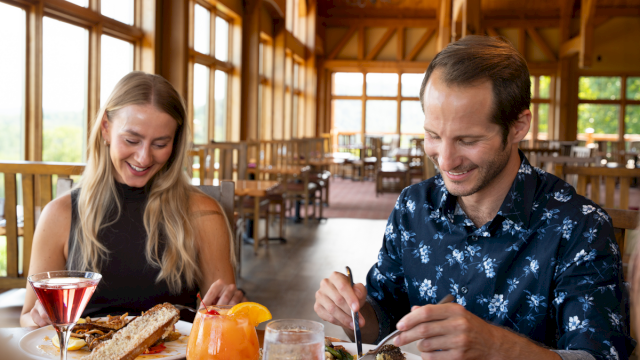 A couple enjoying a bright restaurant meal: woman with wine, man cutting a dish, sunny windowed lodge-style dining, tasty food and orange drinks.