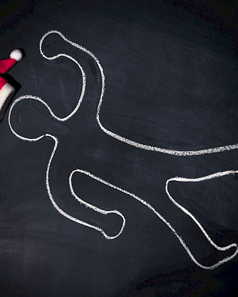 A chalk outline of a person on a dark surface with a Santa hat placed near the head of the outline, suggesting a playful holiday theme.