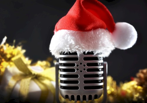 A vintage microphone wearing a Santa hat is in focus, with festive decorations and a gift in the blurred background.