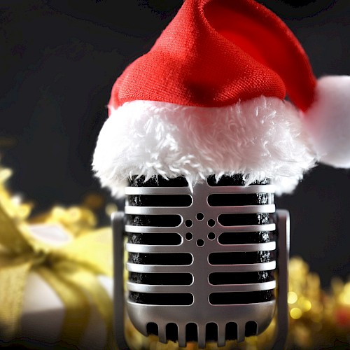 A vintage microphone wearing a Santa hat is in focus, with festive decorations and a gift in the blurred background.