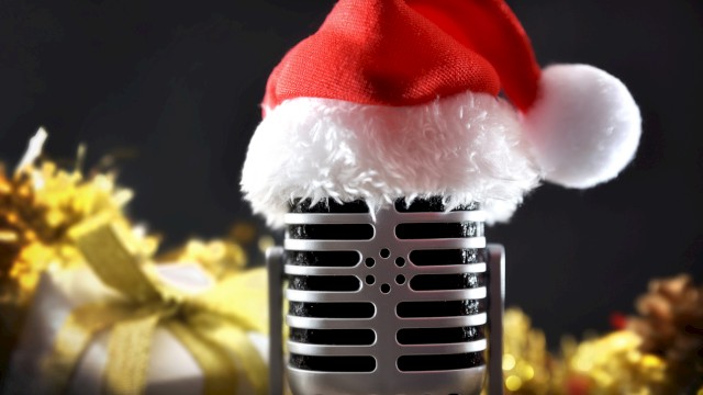 A vintage microphone wearing a Santa hat is in focus, with festive decorations and a gift in the blurred background.