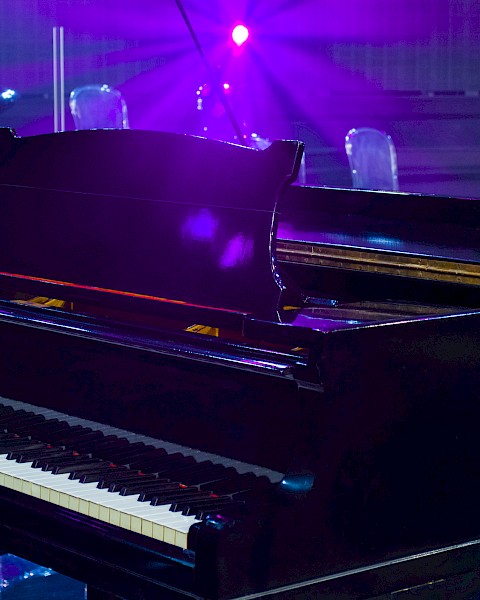A grand piano on a stage with purple lighting and floral decorations in the background.