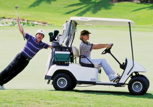 A man drives a golf cart while another clings to the side, playfully holding a golf club on a sunny golf course.
