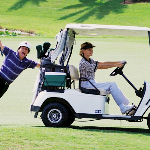A man drives a golf cart while another clings to the side, playfully holding a golf club on a sunny golf course.