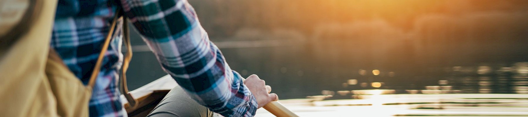 A person in a plaid shirt is canoeing on a calm lake during sunset, holding a paddle, with a backpack visible.