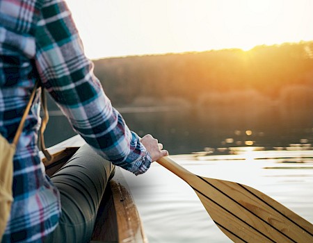 A person in a plaid shirt is canoeing on a calm lake during sunset, holding a paddle, with a backpack visible.