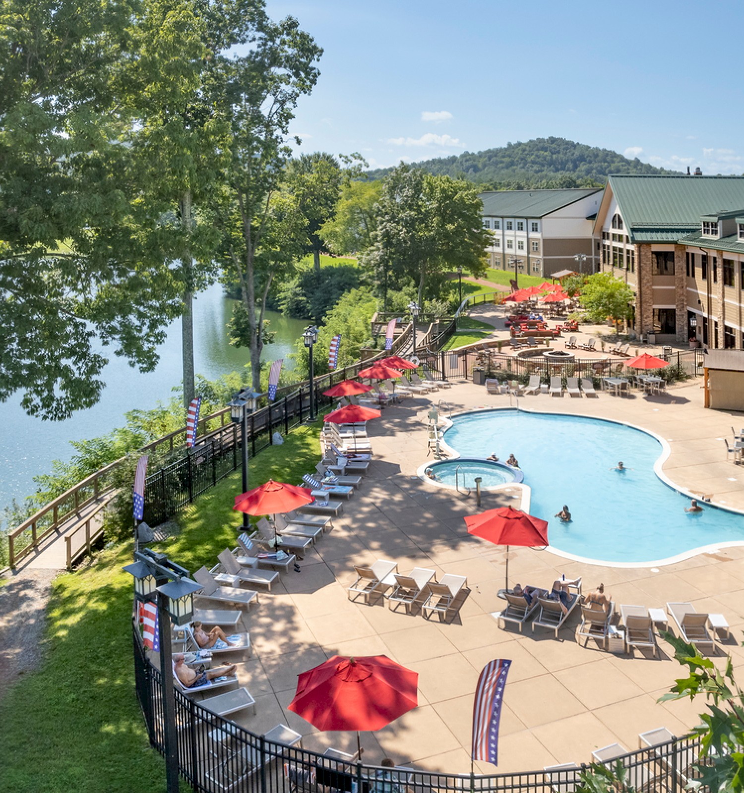 The image shows a scenic resort pool area with red umbrellas, lounge chairs, a lake, lush greenery, and distant hills under a clear blue sky.