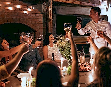 A group of people is gathered around a table, raising glasses in a toast, with candles and food present, in a cozy, dimly-lit setting.
