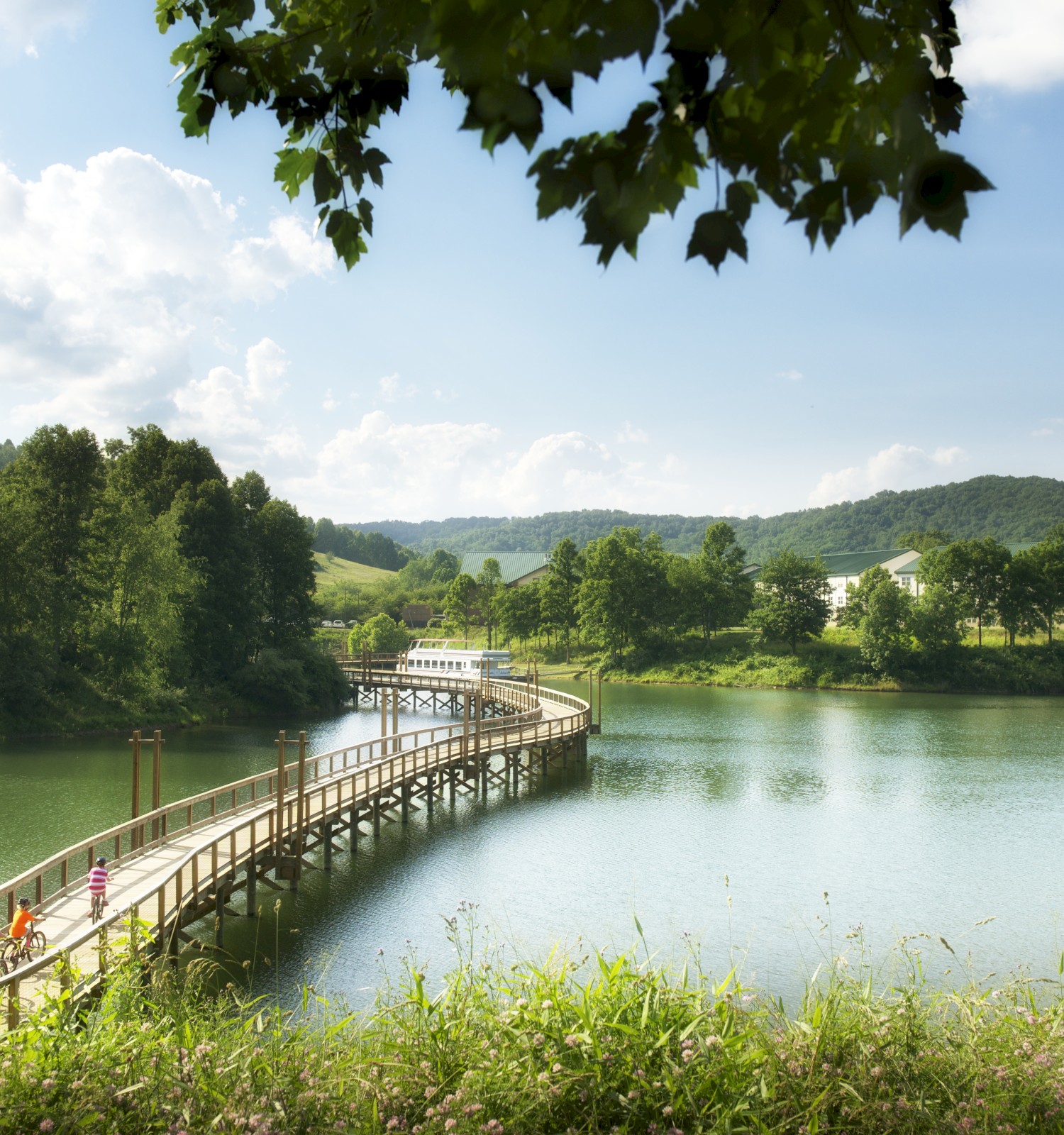A scenic view of a wooden bridge over a tranquil lake surrounded by greenery, with hills and trees under a partly cloudy sky.