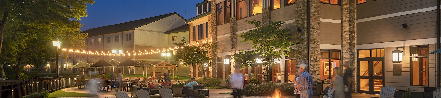 An outdoor patio with a fire pit, seating, string lights, and a building, creating a cozy nighttime atmosphere.