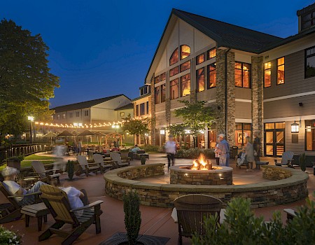 An outdoor patio with a fire pit, seating, string lights, and a building, creating a cozy nighttime atmosphere.