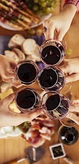 A group of people clinking glasses of red wine over a table with food, including bread, cheese, and meats, in a celebratory setting.