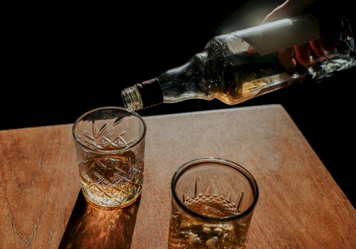 A hand pours amber liquid from a bottle into two cut-glass tumblers on a wooden surface, against a dark background.