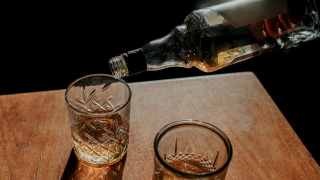 A hand pours amber liquid from a bottle into two cut-glass tumblers on a wooden surface, against a dark background.
