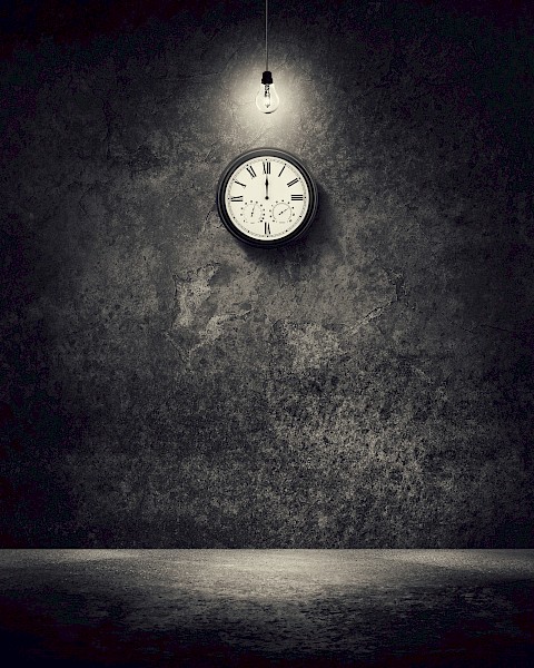 A dimly lit room with a hanging lightbulb and a wall clock showing 10:10, casting shadows on a textured wall.