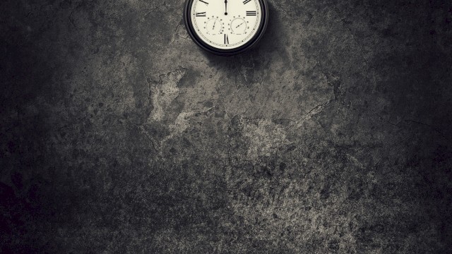 A dimly lit room with a hanging lightbulb and a wall clock showing 10:10, casting shadows on a textured wall.
