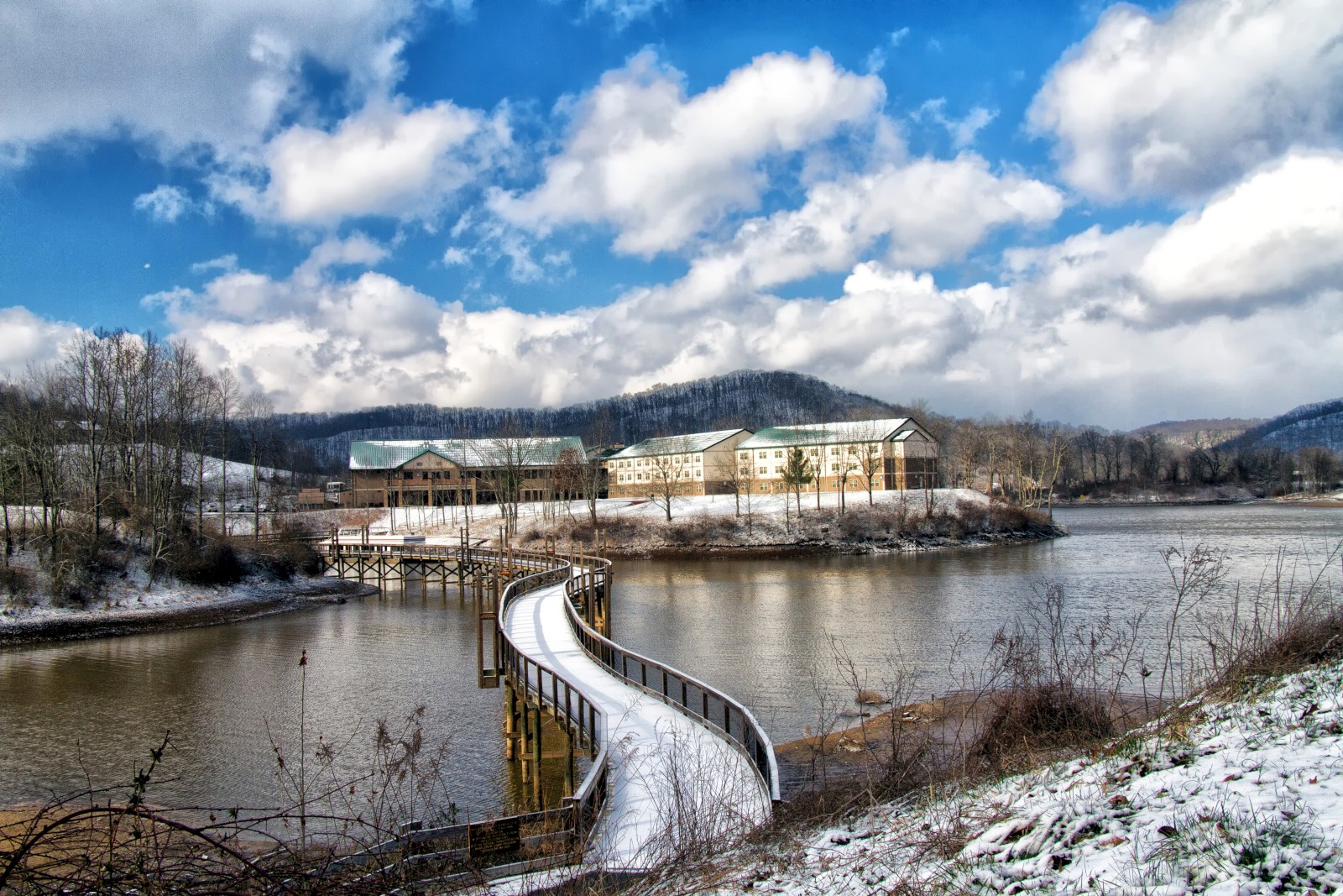 A snowy riverside park with a curved boardwalk leading to a lakeside building, under a bright blue sky with fluffy clouds.