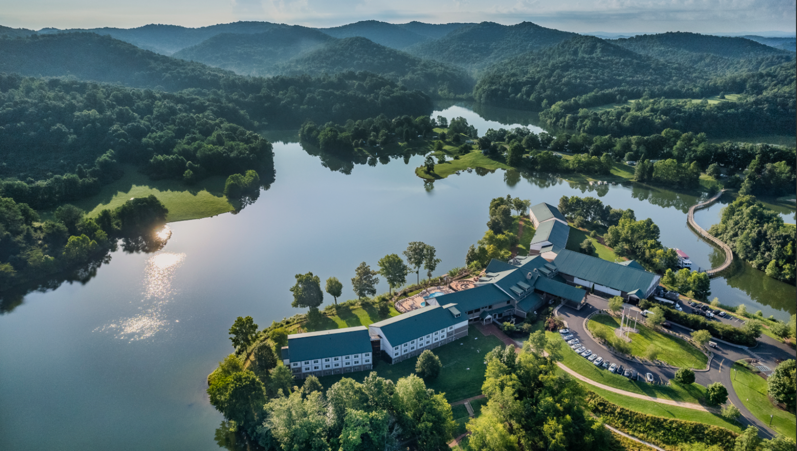 Aerial view of buildings surrounded by a lake, trees, and hills under a cloudy sky, capturing a serene and scenic landscape.