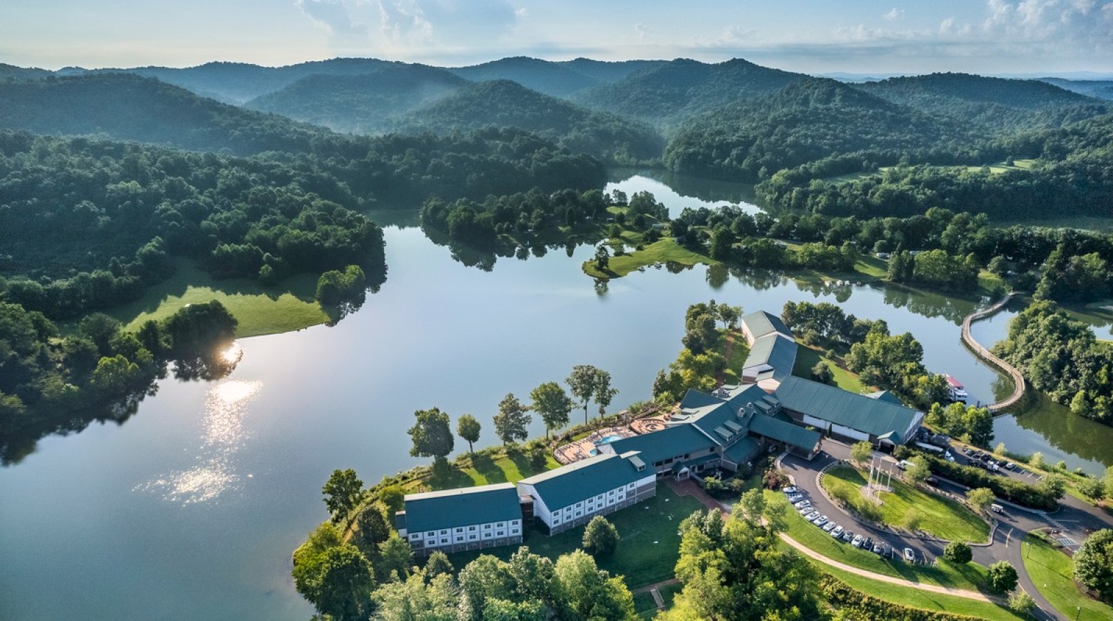 Aerial view of a lush, forested landscape with a large building by a serene lake, surrounded by rolling hills under a clear sky.