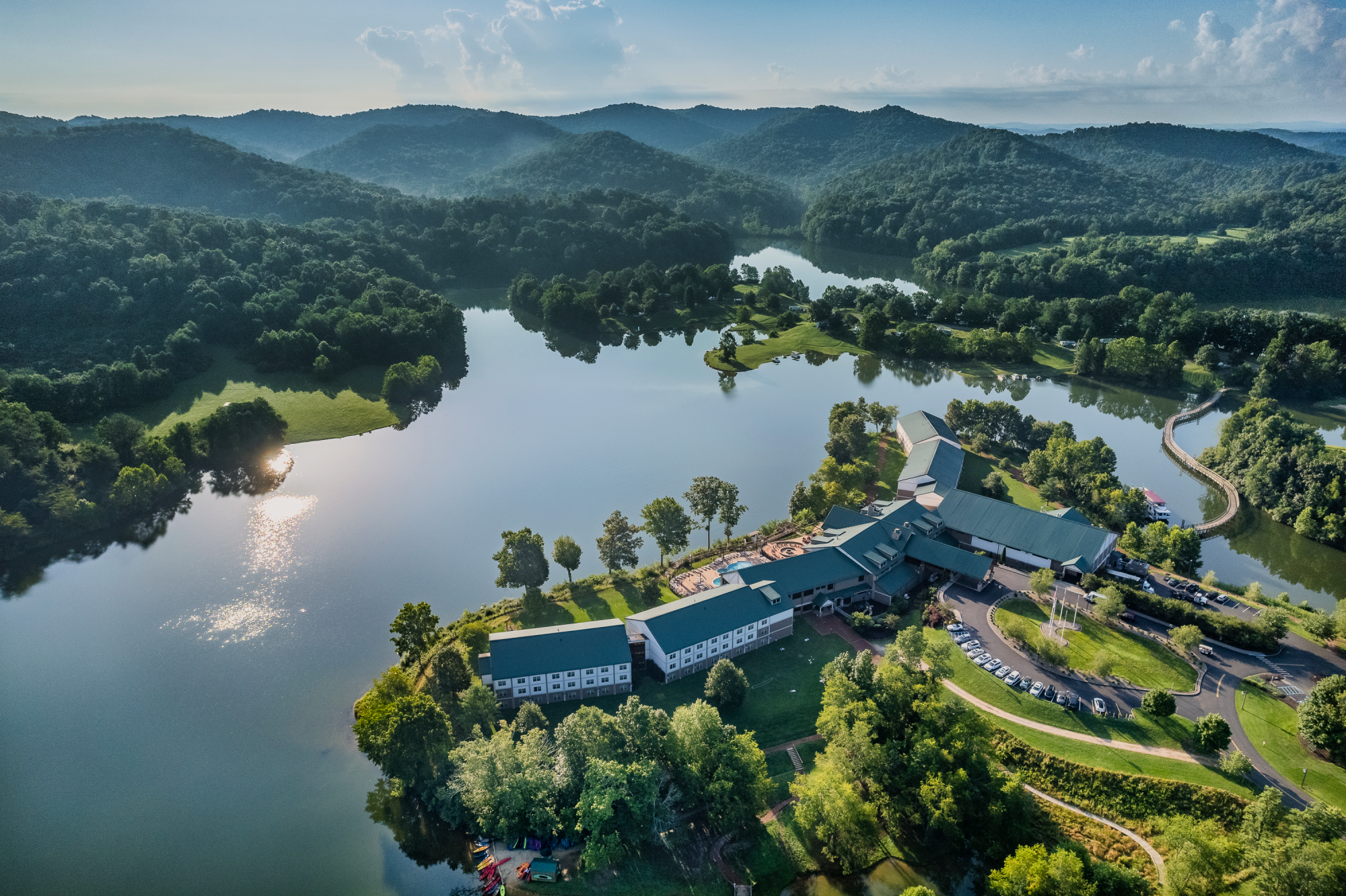 An aerial view of a serene lakeside resort surrounded by lush green hills and trees, with clear blue skies above and sunlight reflecting off the water.