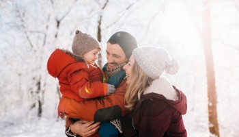 A family of three in a snowy forest: a mother and father smile at their toddler bundled in a red snowsuit, warm hats, and cozy scarves.