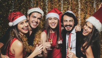 A group of five friends in festive outfits—Santa hats, red dresses, and a suit—pose with champagne glasses, smiling against a glittery backdrop.