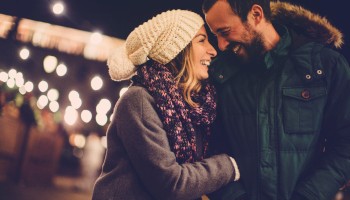 Two people smiling warmly at a cozy outdoor night scene, bundled in winter coats, scarves, and a knit hat, sharing an affectionate moment.
