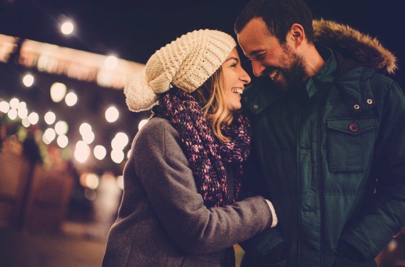 Two people smiling warmly at a cozy outdoor night scene, bundled in winter coats, scarves, and a knit hat, sharing an affectionate moment.