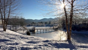 A snowy landscape with a sunlit tree, a wooden bridge over a frozen lake, and mountains in the background.