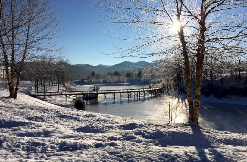 A snowy landscape with a sunlit tree, a wooden bridge over a frozen lake, and mountains in the background.