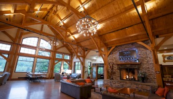 A spacious room with wooden beams, large windows, comfortable seating, and a stone fireplace under a chandelier.